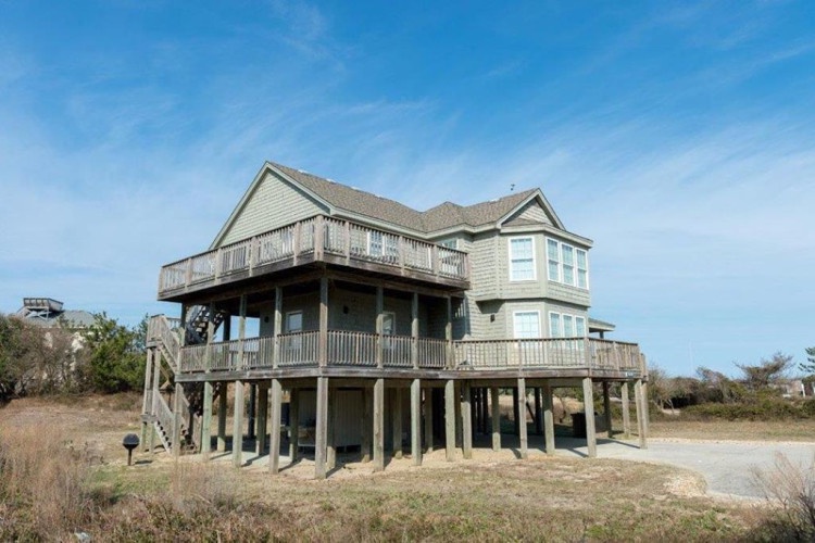 Elevated coastal vacation home on stilts with wraparound decks, offering sweeping ocean views and beach access in a natural dune setting.