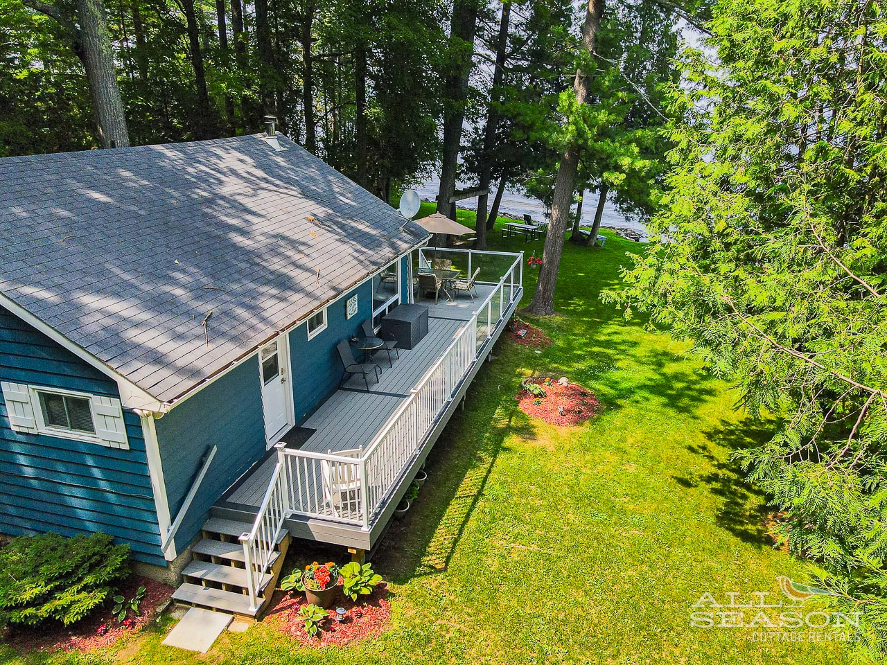 Aerial view of charming blue cottage with spacious deck surrounded by mature trees and lawn.