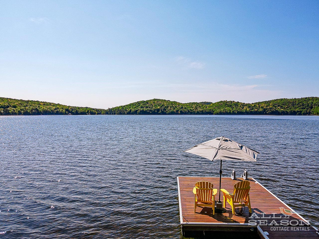 Peaceful lake setting with rolling hills, featuring a private dock with yellow chairs and umbrella for relaxing waterside moments.