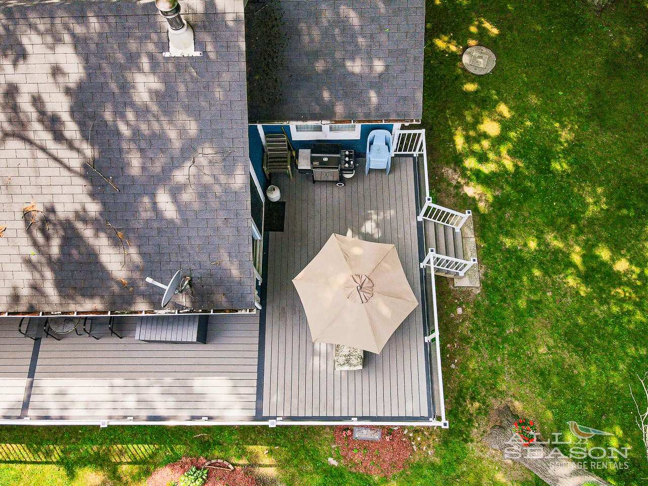 Aerial view of the property's outdoor deck with BBQ facilities, patio umbrella, and expansive lawn area surrounded by mature trees.