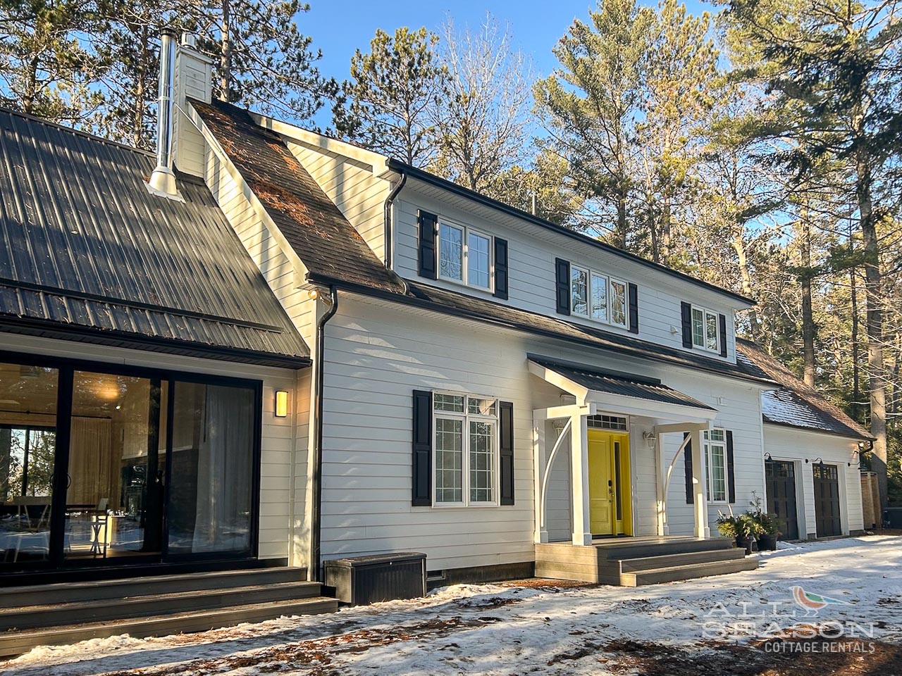 Modern white cottage nestled among towering pines with welcoming yellow door and snow-dusted entrance.