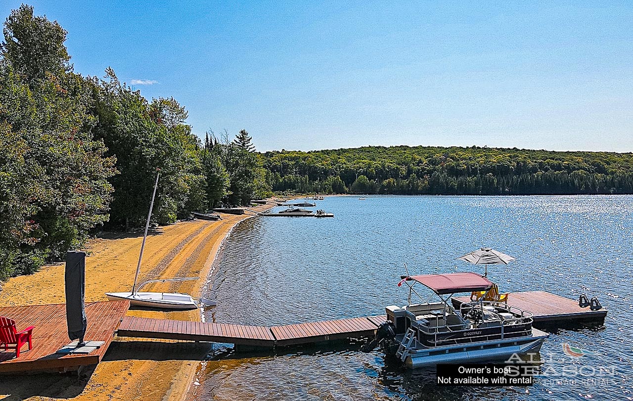 Sandy lakefront with private dock and pontoon boat surrounded by forested shoreline under clear blue skies.