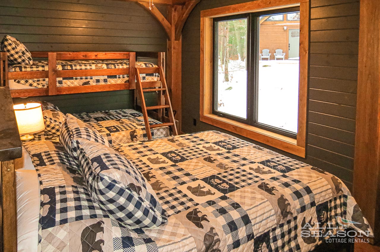 Cozy up in this rustic bunk room with plaid bedding and timber beams, where snowy winter views create your perfect cabin retreat.