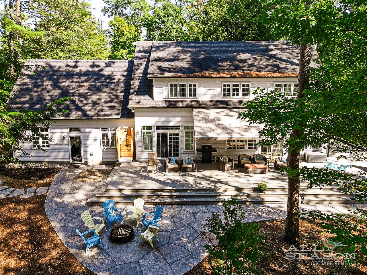 Spacious lakefront cottage surrounded by mature trees, featuring multiple outdoor seating areas and pristine natural setting.