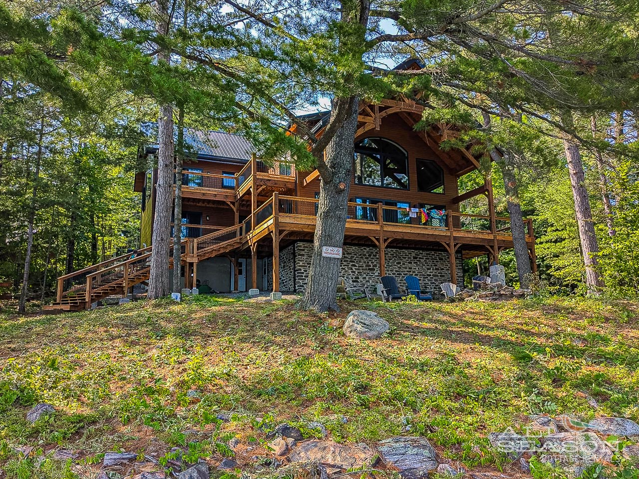 Stunning lakeside cabin nestled among towering trees, featuring multi-level decks and rustic stone foundation in peaceful woodland setting.