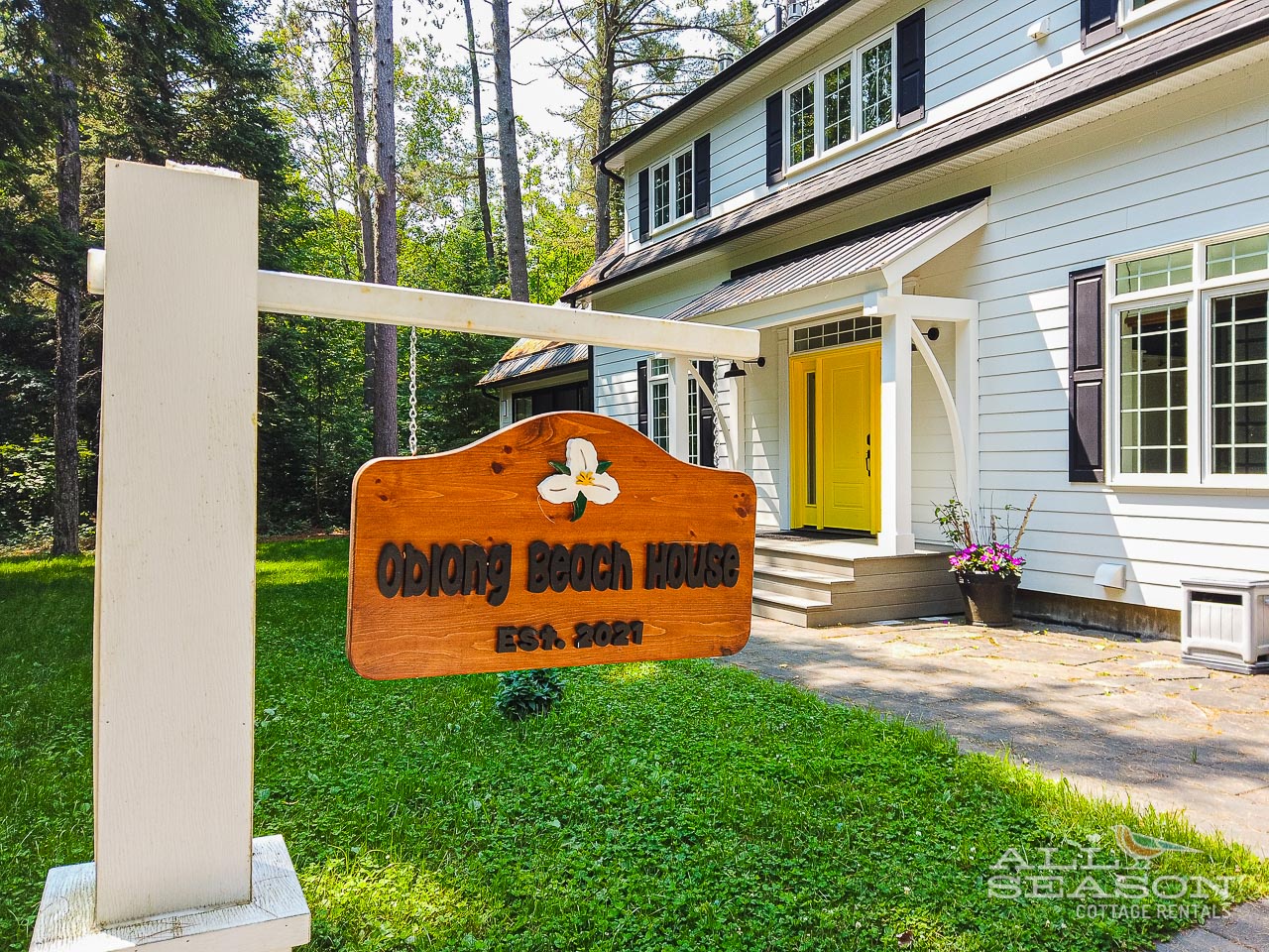 Welcome to Oblong Beach House, established 2020. A charming cottage retreat nestled among towering trees with a cheerful yellow door welcoming your arrival.