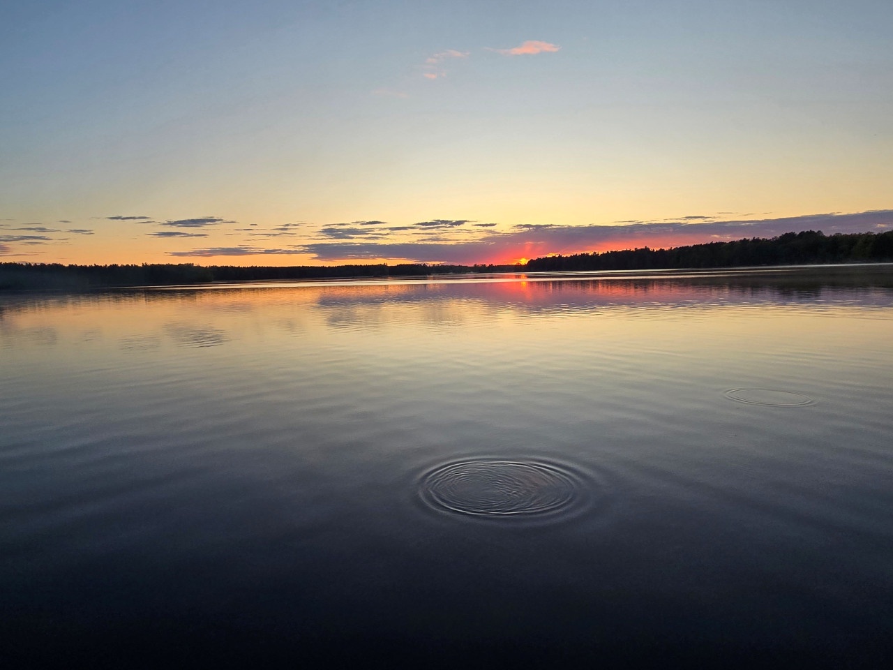 Tranquil lake at sunset with gentle ripples across calm waters, forested shoreline creating a peaceful natural setting.