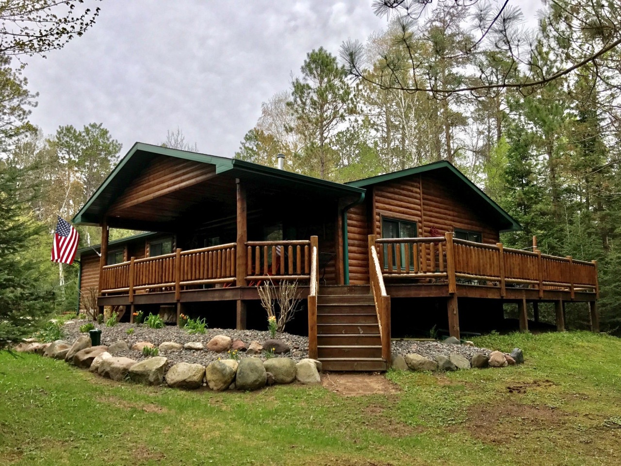 Charming log cabin nestled among towering trees with spacious wraparound deck featuring a covered porch and American flag.