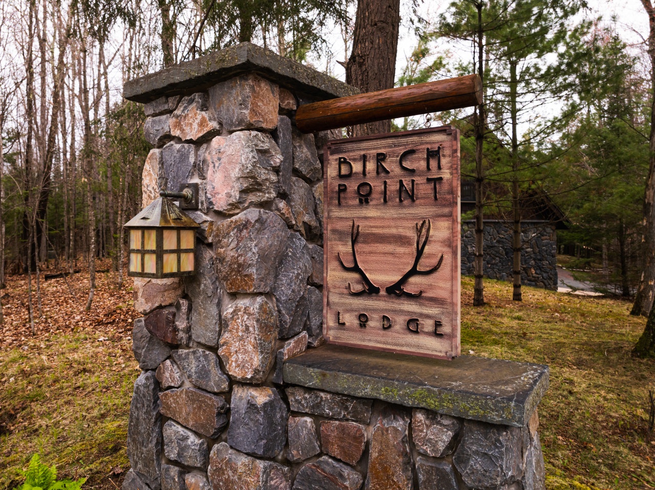 Stone entrance marker with rustic wooden sign welcomes guests to Birch Point Lodge, nestled among towering trees and natural woodland setting.