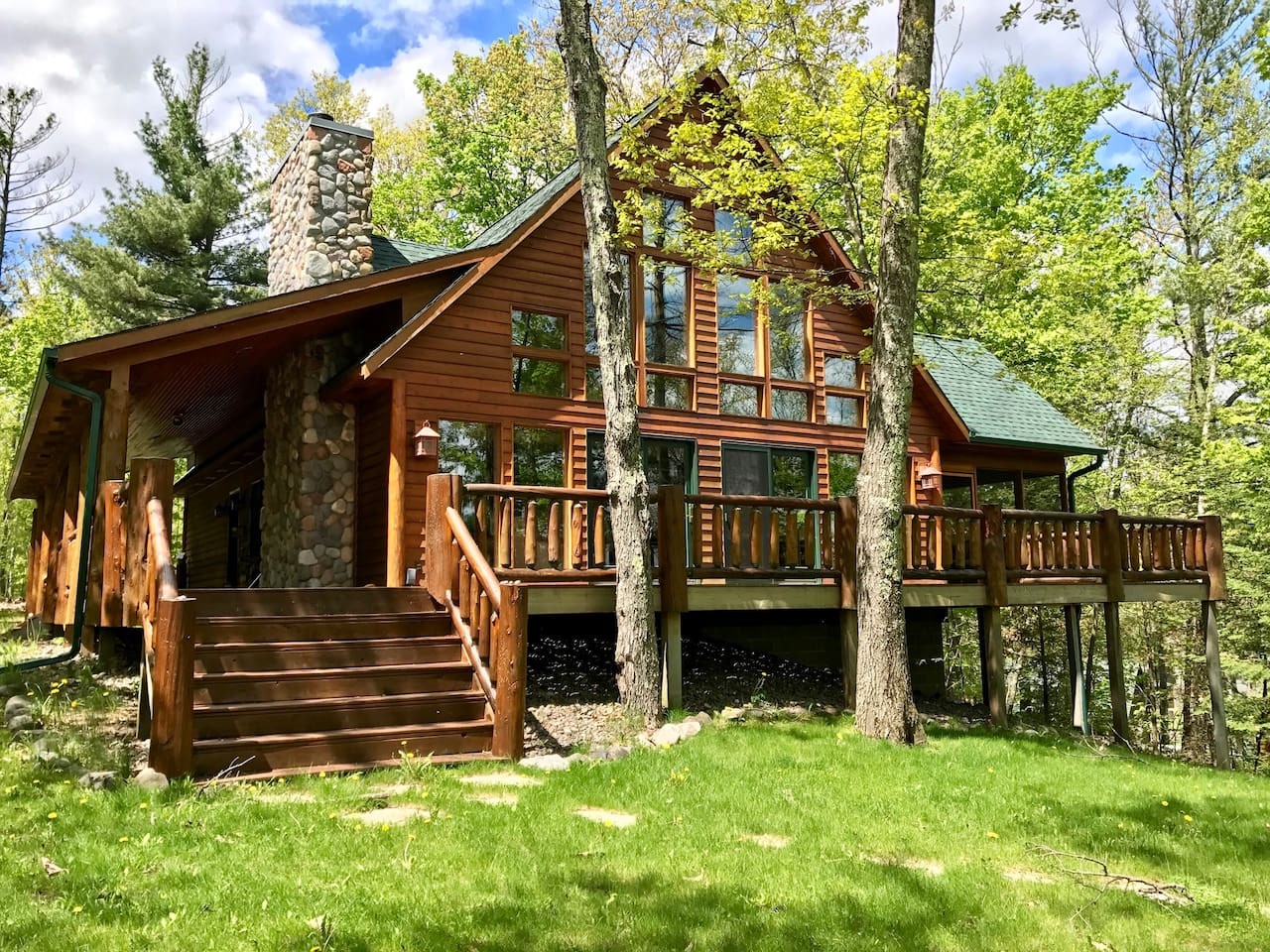 A beautiful log cabin nestled among towering trees features stone accents and wraparound decks for enjoying the peaceful forest setting.