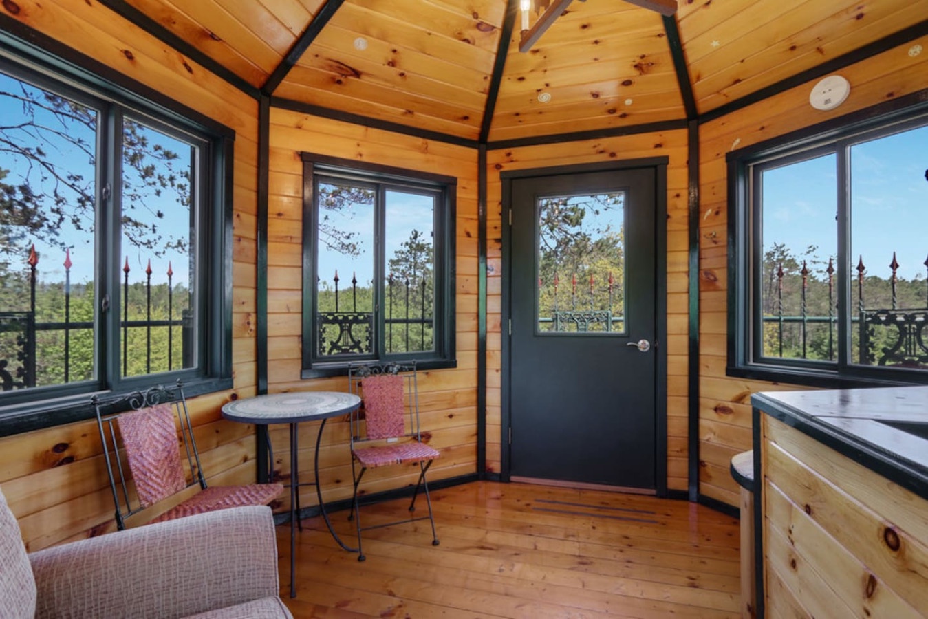 Relax in your private wood-paneled sunroom with panoramic windows showcasing forest views through ornate iron railings.