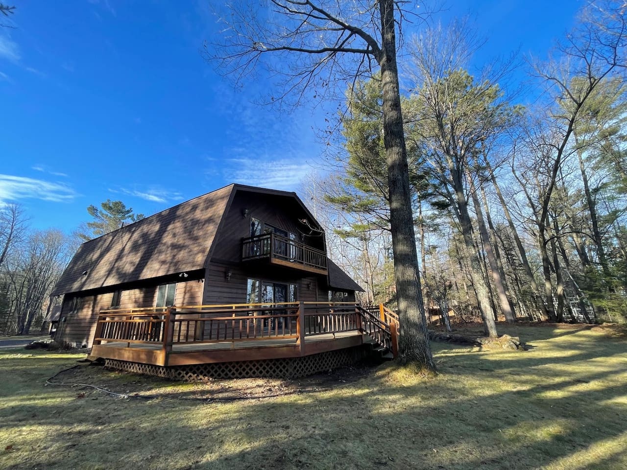 A charming woodland cabin featuring wraparound decks and rustic charm, surrounded by towering trees on a sunny spring day.