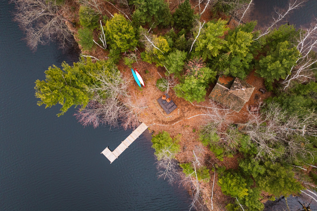 Aerial view of a secluded waterfront property featuring private dock, kayaks, and fire pit surrounded by natural forest landscape.