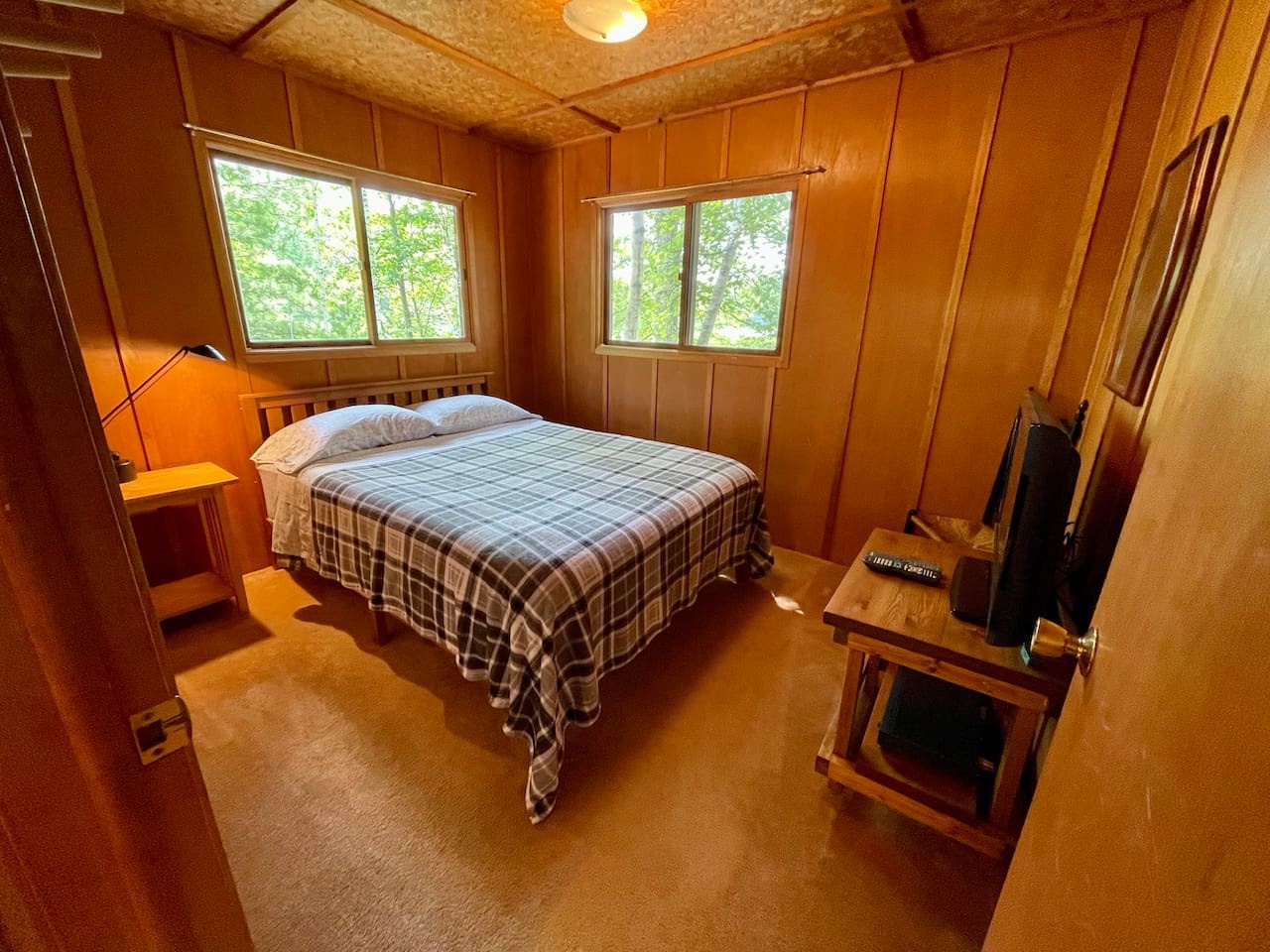 Unwind in your cozy wood-paneled bedroom featuring warm plaid bedding and peaceful forest views through your windows.