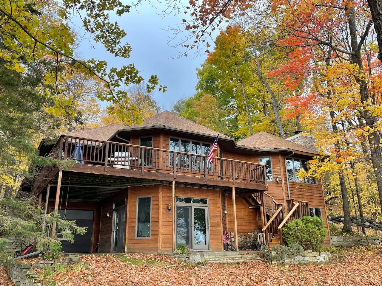 A beautiful two-story wooden cabin nestled among vibrant autumn trees, featuring wraparound decks and peaceful forest surroundings.