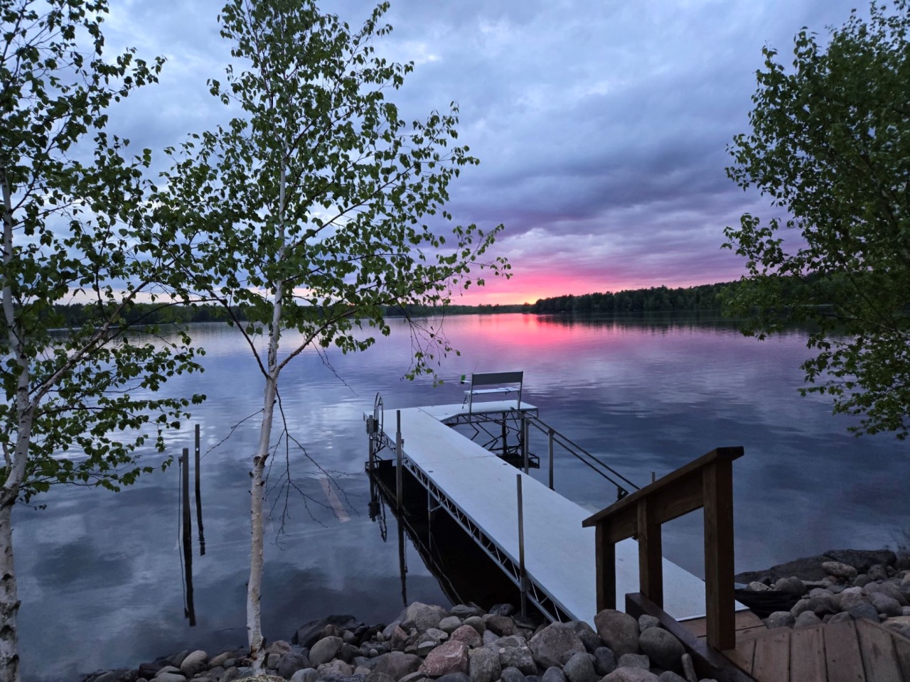A stunning lakeside sunset paints the sky in vibrant pink and purple hues, with a peaceful dock extending over calm waters.