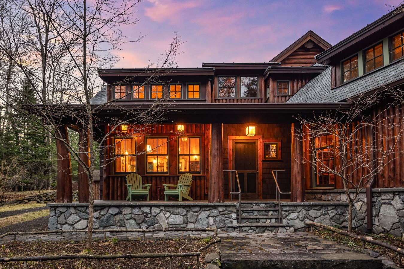 Rustic mountain lodge exterior featuring warm wood siding and stone foundation, illuminated windows creating a welcoming glow at twilight.