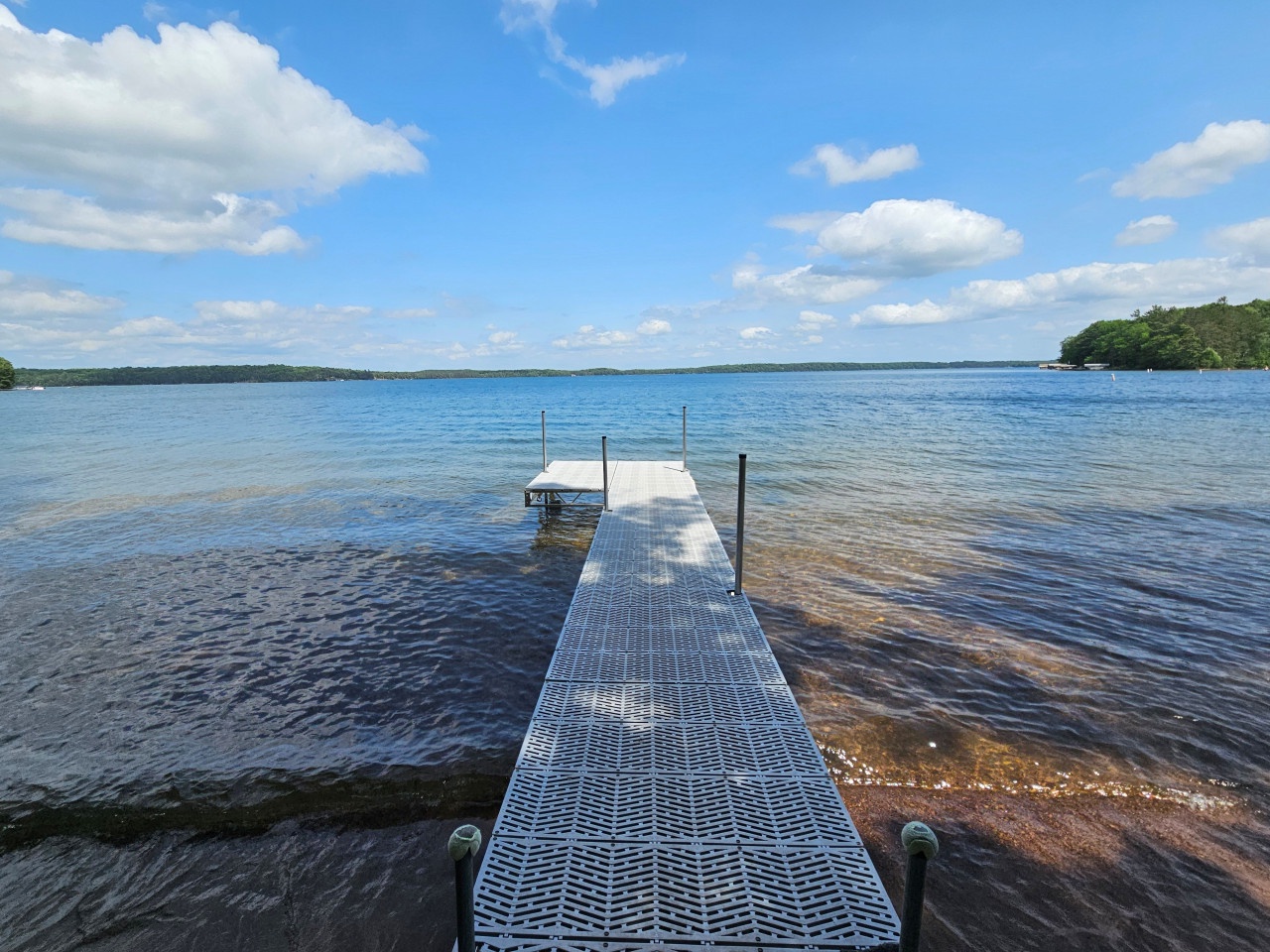 Private dock extending into pristine lake waters, surrounded by forested shoreline under expansive blue skies with white clouds.