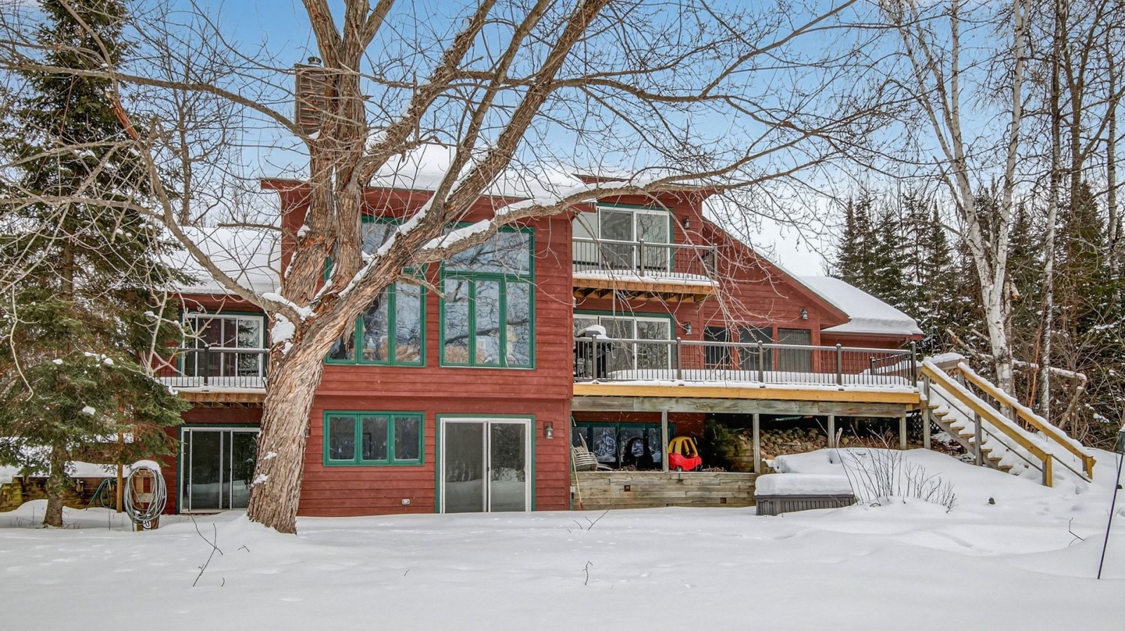 A charming red lakefront property surrounded by pristine snow and mature trees, featuring multiple levels and extensive outdoor decking.