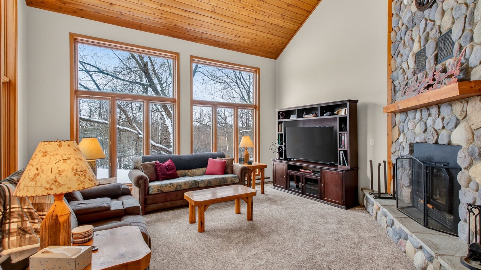 Relax in your cozy living room with soaring wood ceilings, stone fireplace, and large windows overlooking peaceful winter woods.