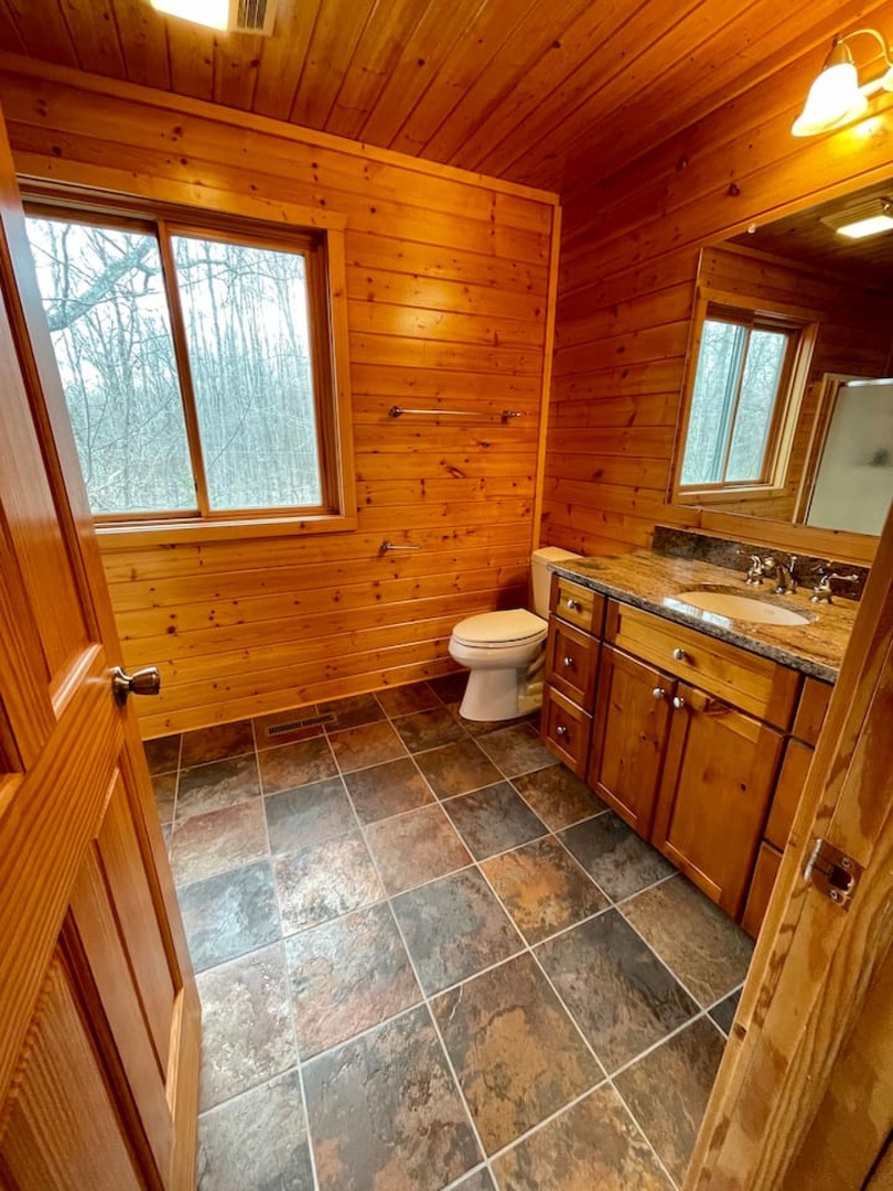 Relax in your rustic bathroom with warm wood-paneled walls and slate floors, where forest views from the window create a peaceful retreat.