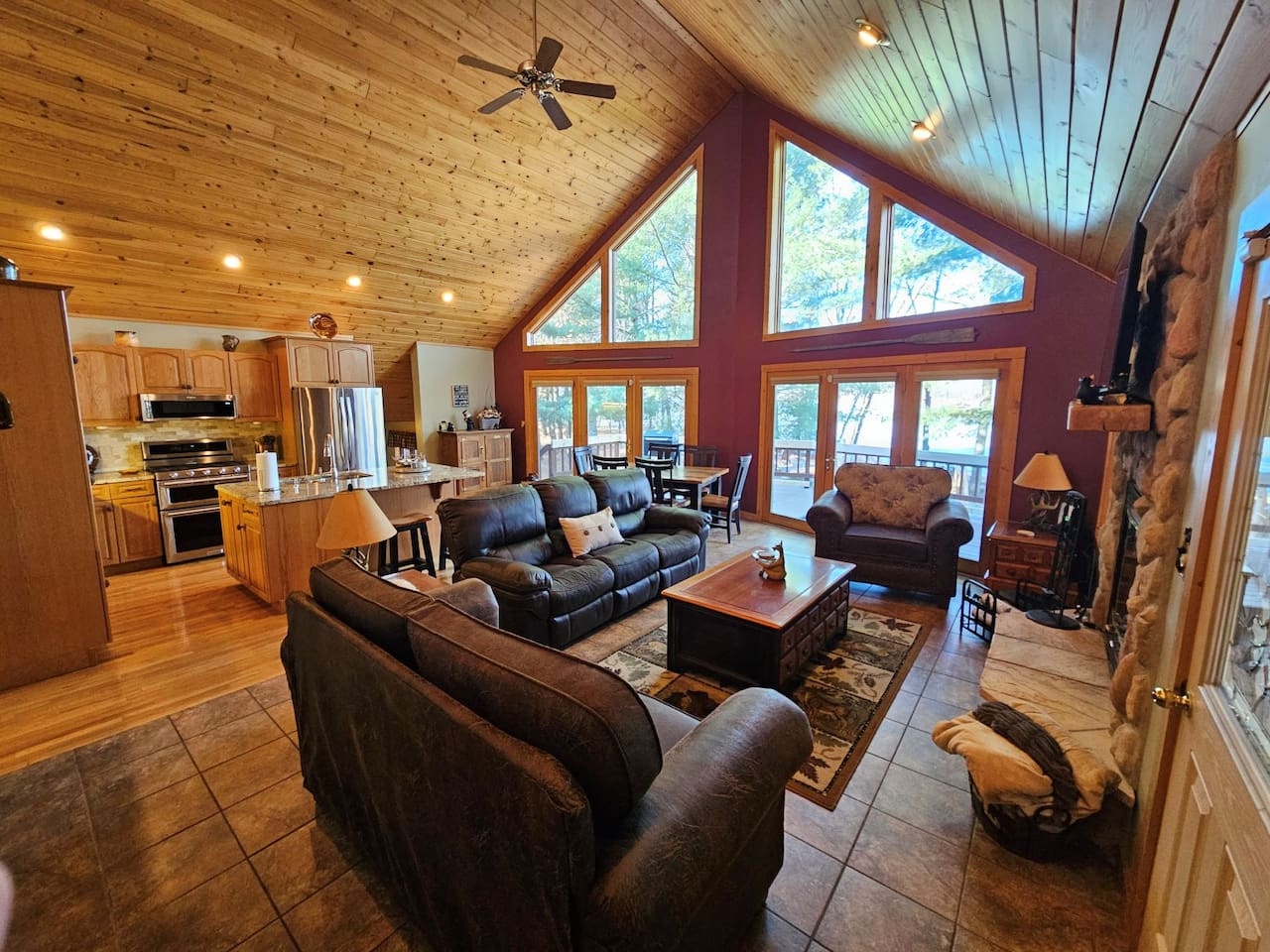 Unwind in your cozy cabin living space with soaring ceilings, a stone fireplace, and panoramic windows framing forest views.