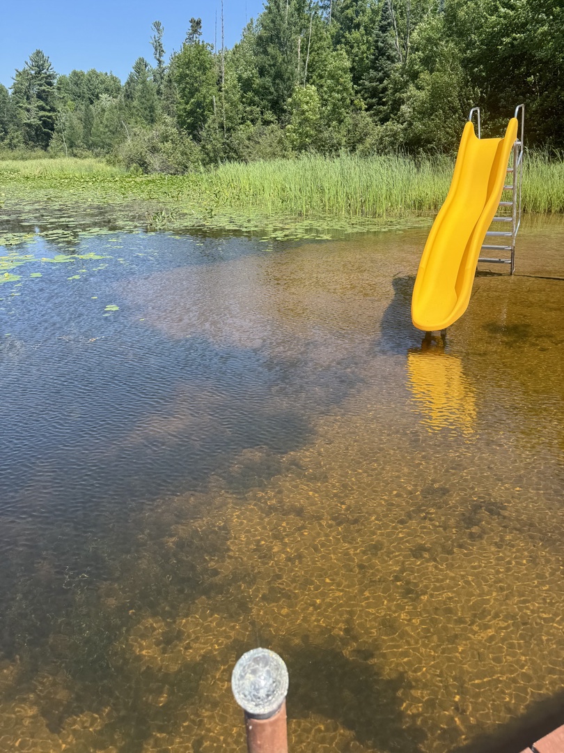 A peaceful lakeside area with a bright yellow slide positioned in calm, shallow waters surrounded by lush greenery.