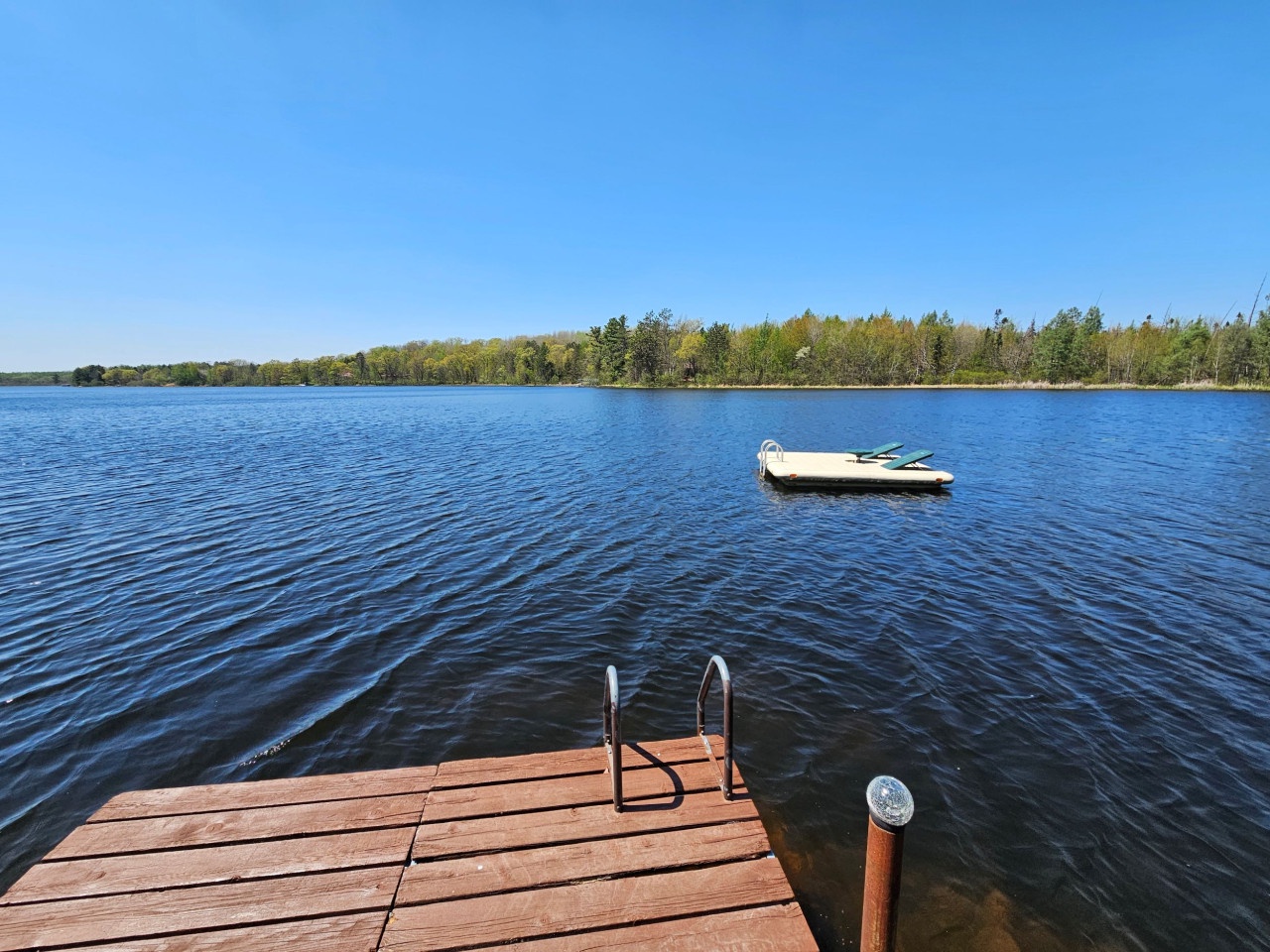Pristine lake with wooden dock and boat anchored nearby, surrounded by lush forest under clear blue skies.