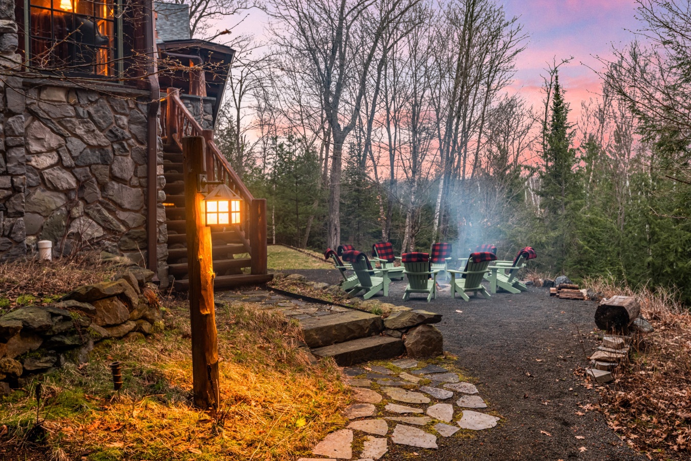 Rustic stone cottage surrounded by forest with Adirondack chairs around a fire pit at twilight.