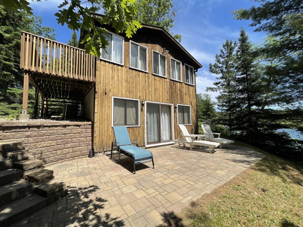 Charming lakefront cabin featuring natural wood siding, expansive windows, and elevated deck surrounded by lush forest.