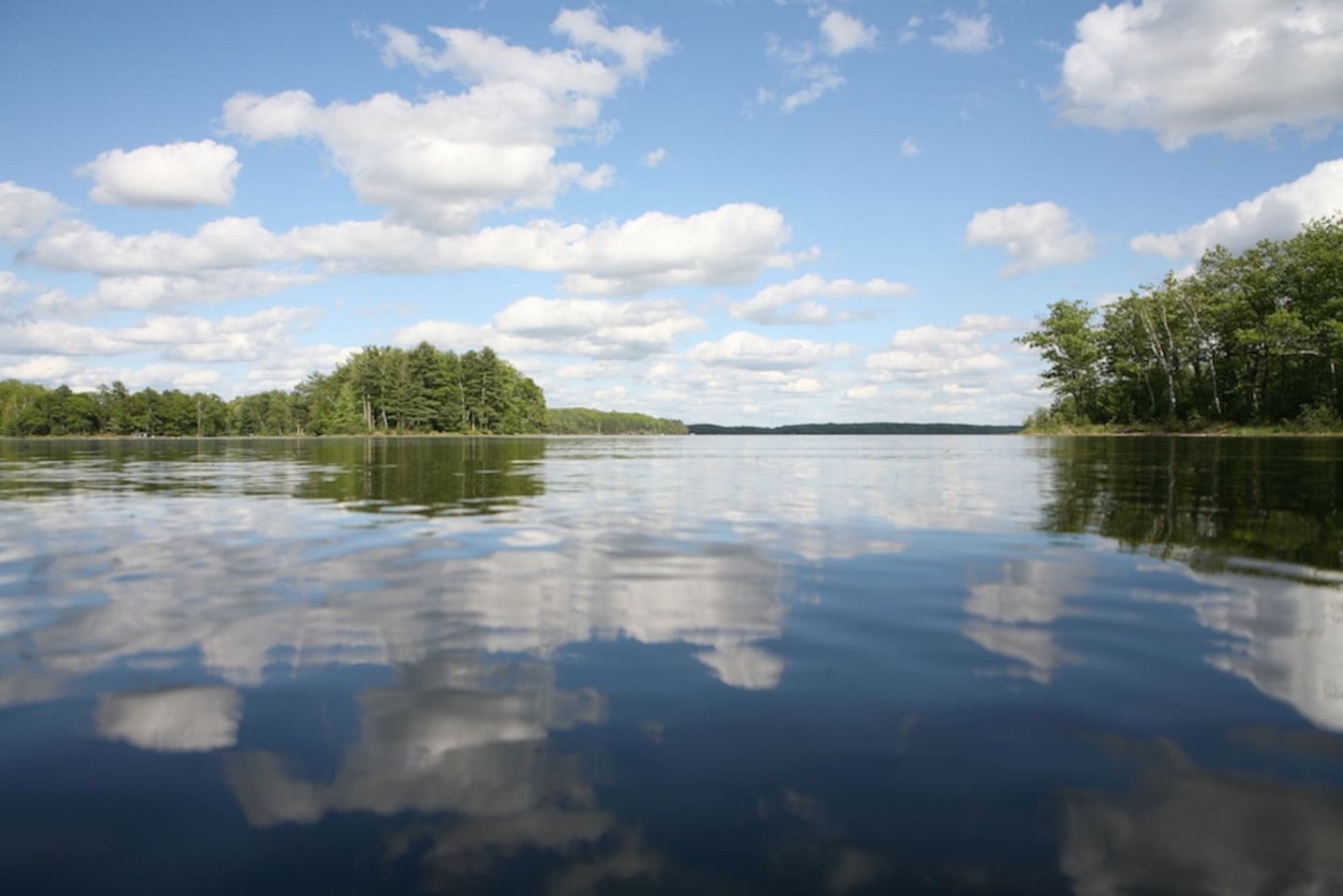 Serene lake reflects puffy clouds and lush forests, creating perfect mirror-like views from this peaceful waterfront location.