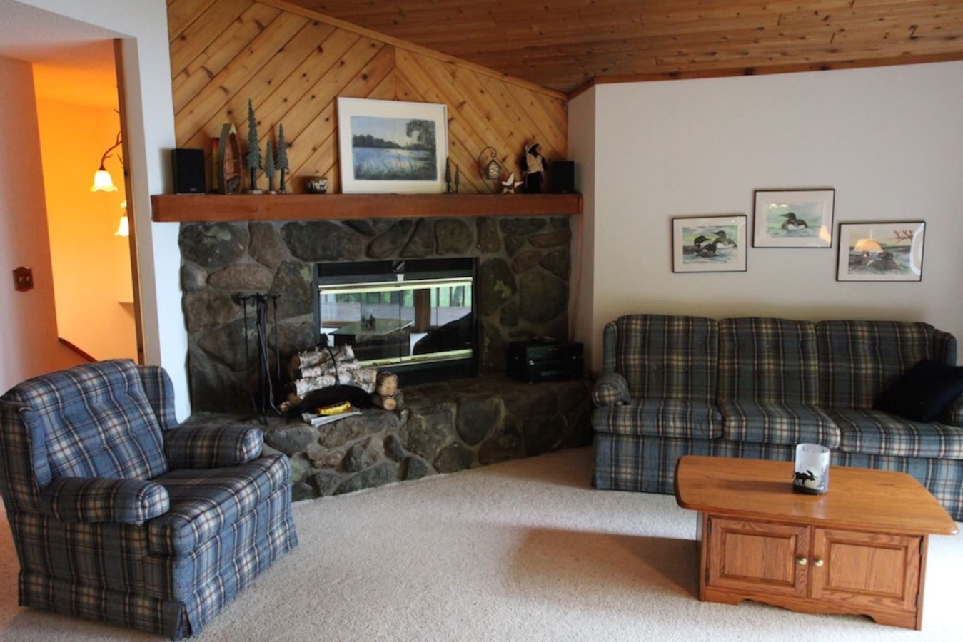 Unwind in your cozy living room featuring a stunning stone fireplace, plaid furnishings, and warm wood accents that create the perfect lakeside retreat atmosphere.