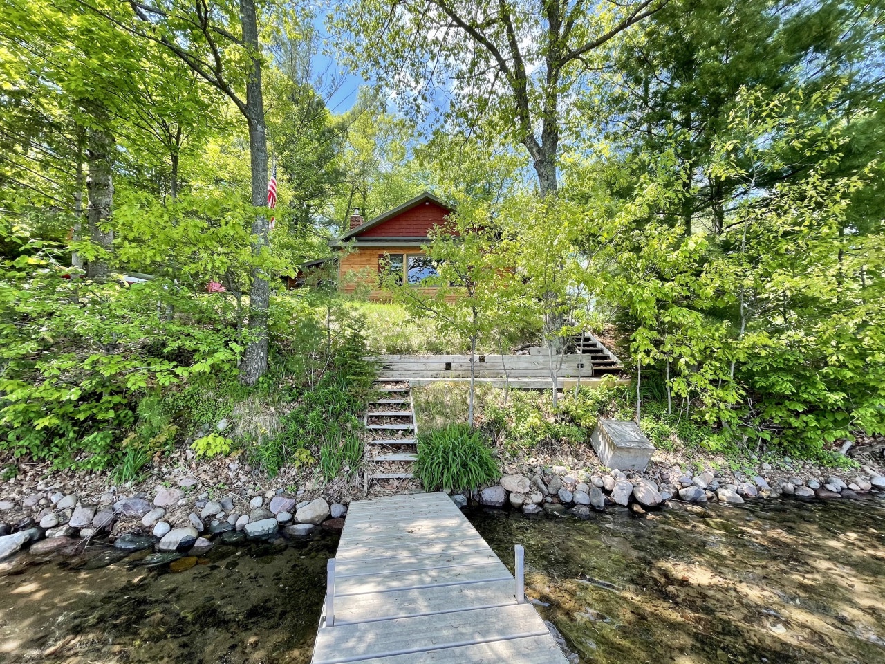 Lakefront cabin nestled among towering trees with private dock and stone pathways leading through lush natural surroundings.