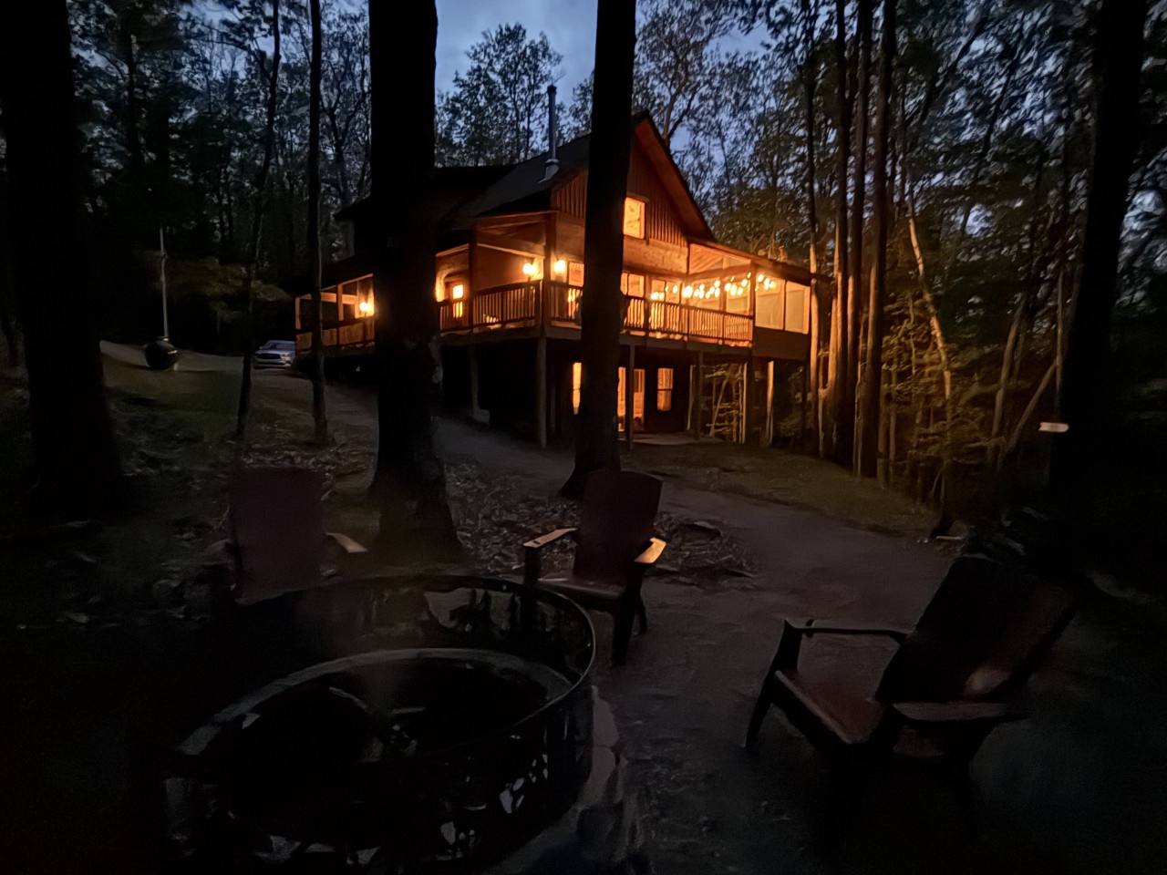 Secluded forest cabin glows warmly at twilight, featuring outdoor seating and fire pit surrounded by towering trees.