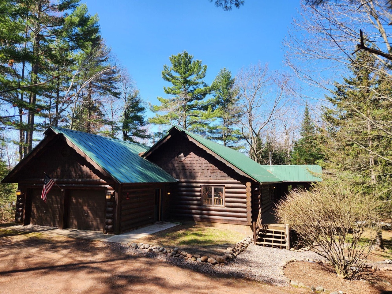 Rustic log cabin nestled among towering pines with green metal roofing and natural stone landscaping in a peaceful forest setting.