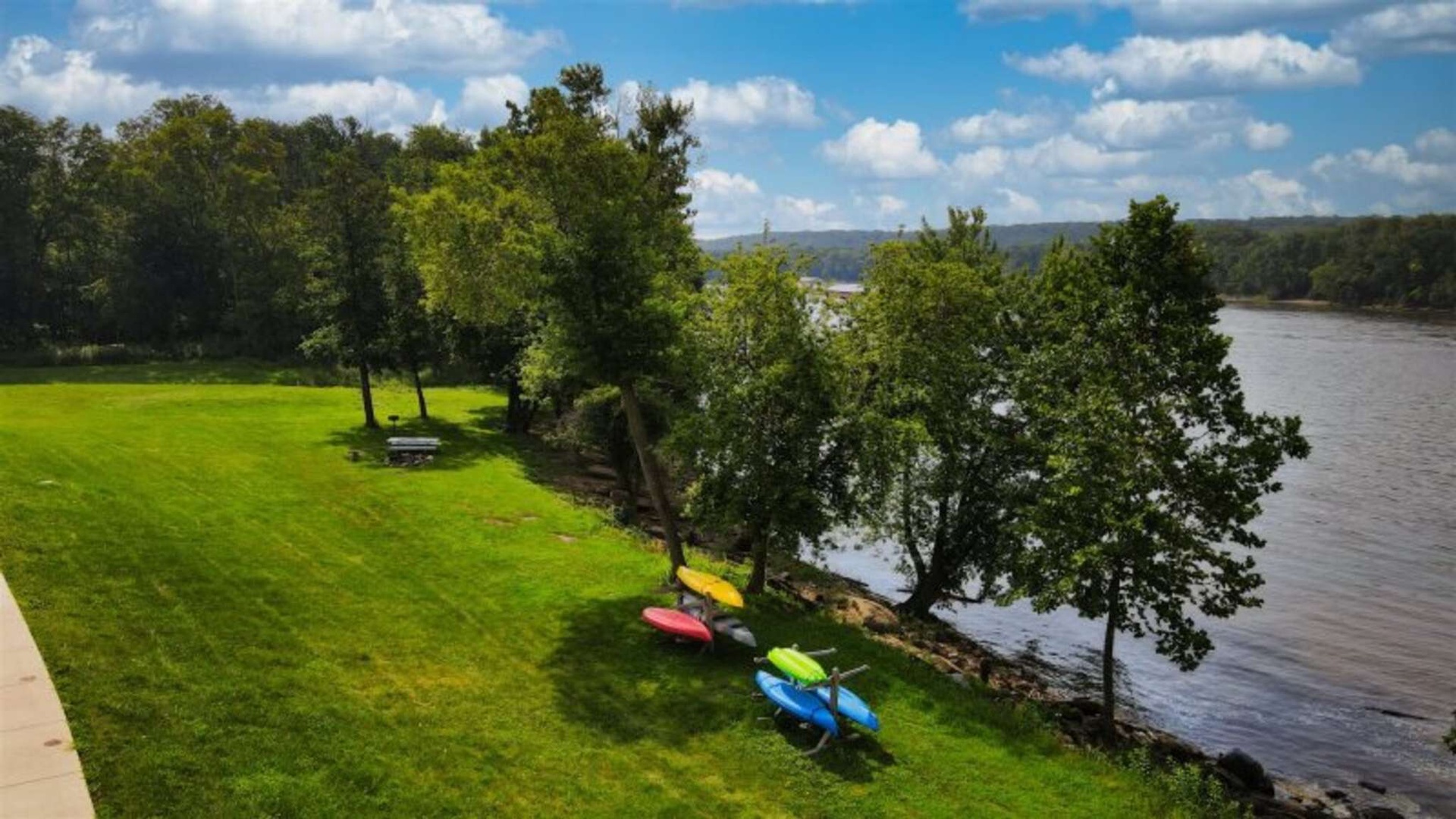Scenic waterfront property features colorful kayaks on pristine lawn beside a peaceful river, surrounded by mature trees and rolling hills.