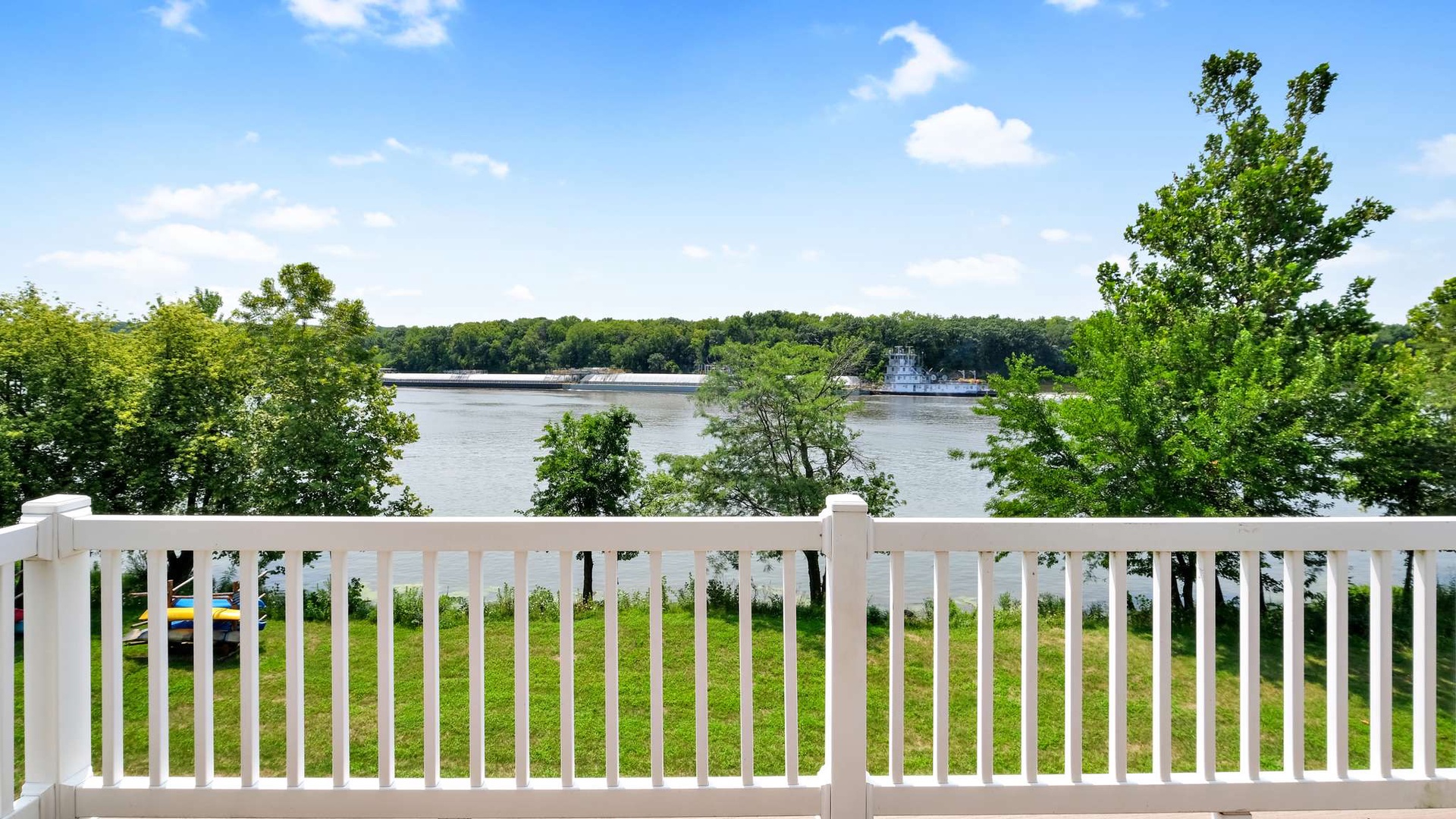 Peaceful lake views stretch beyond the property's white railing, surrounded by lush trees under bright blue skies.