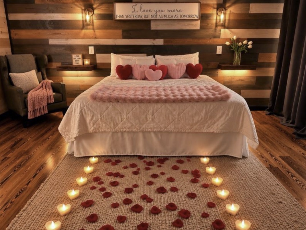Settle into your romantic retreat, where rose petals and candlelight frame a cozy bed beneath warm wood tones and a loving message overhead.