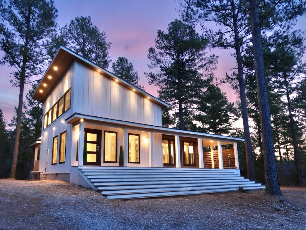 A modern barn-style retreat glows warmly at dusk, nestled among tall pines with a painted sky behind. The wide front steps and wrap-around deck invite a peaceful arrival in nature.