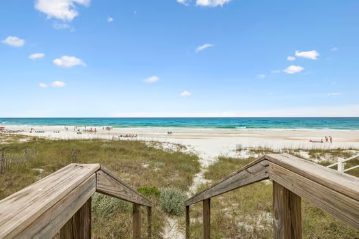 Wooden boardwalk steps lead to pristine white sand beaches and turquoise Gulf waters, with gentle dunes and scattered beachgoers visible along the shore.