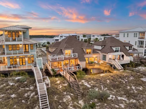 These beachfront vacation homes feature elevated designs with stunning water access via private boardwalks leading directly to the sandy shore, with warm lighting welcoming guests at sunset.