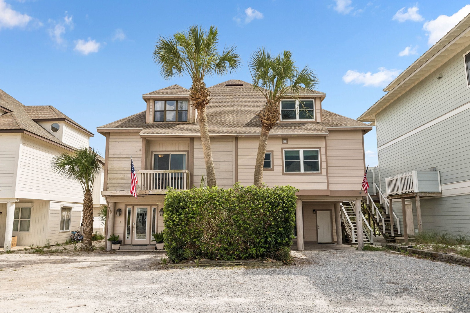 A welcoming coastal property featuring a three-story beachside home with palm trees, ground-level parking, and a comfortable front porch balcony area.