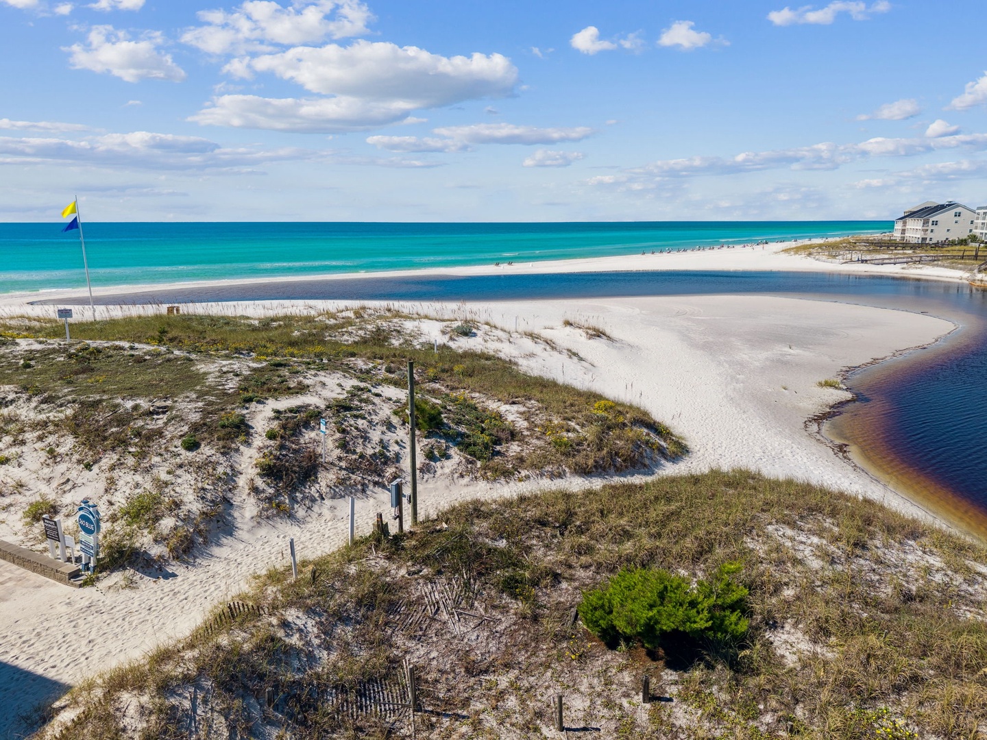 An aerial view captures the pristine white sands and turquoise waters just steps from the property, with coastal dunes and nearby buildings visible along the shore.