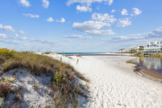 Pristine white sand beach bordered by natural dunes and a peaceful waterway, with coastal homes visible in the distance under a clear blue sky.