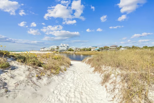 A sandy coastal path winds through native dune grasses toward a cluster of beachfront homes beneath a bright blue sky dotted with scattered clouds.