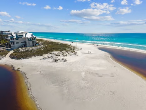 Aerial view of the property's beachfront location featuring pristine white sand, turquoise Gulf waters, and coastal dune vegetation. Nearby vacation homes are visible along the shore.