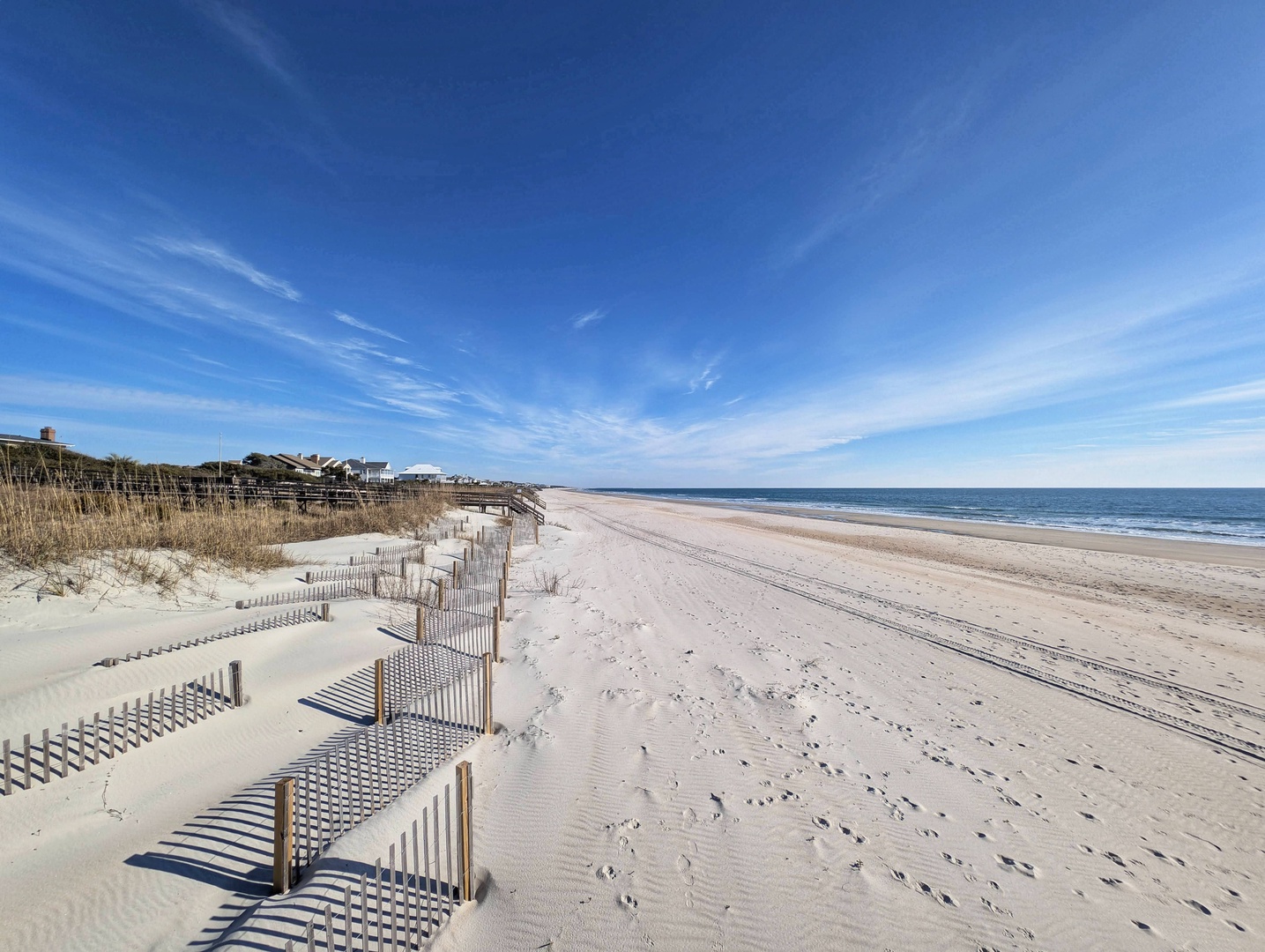 Expansive white sand beach stretches along pristine shoreline, bordered by natural dunes and calm blue ocean waters under clear skies.