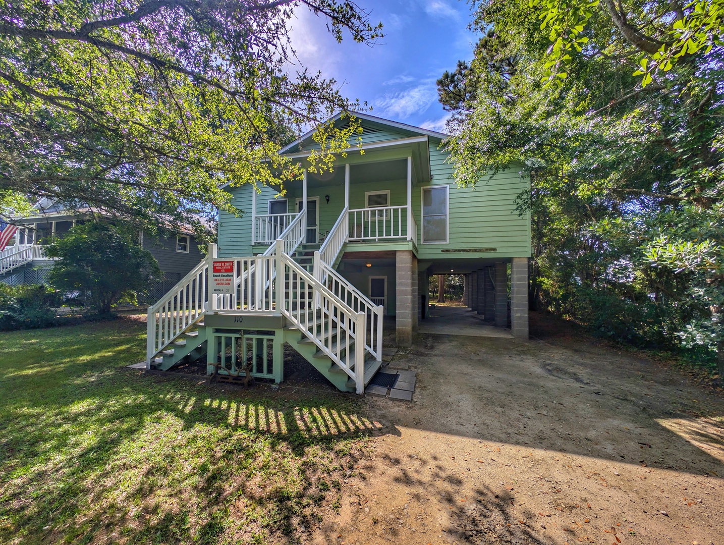 A charming raised beach cottage framed by mature trees offers covered ground-level parking and welcoming front stairs leading to the main living space, nestled in a peaceful neighborhood setting.