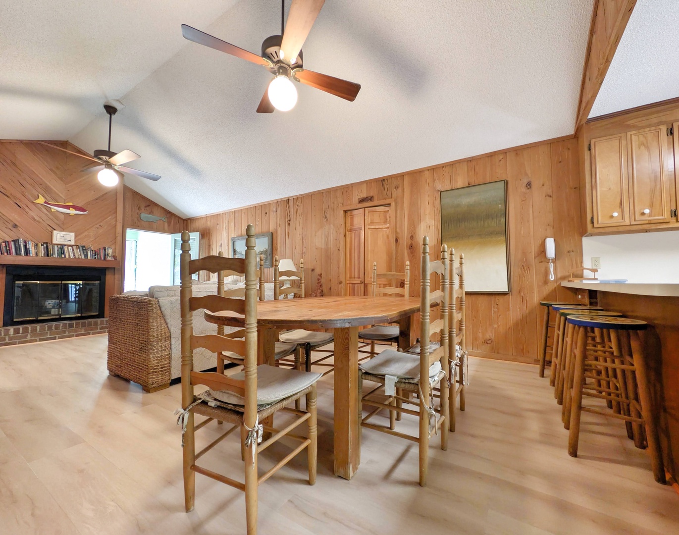 Gather around the handcrafted wooden dining table in this warm, pine-paneled space with vaulted ceilings and ceiling fans. The adjacent breakfast bar offers extra seating, while the cozy fireplace in the living area creates a welcoming retreat.