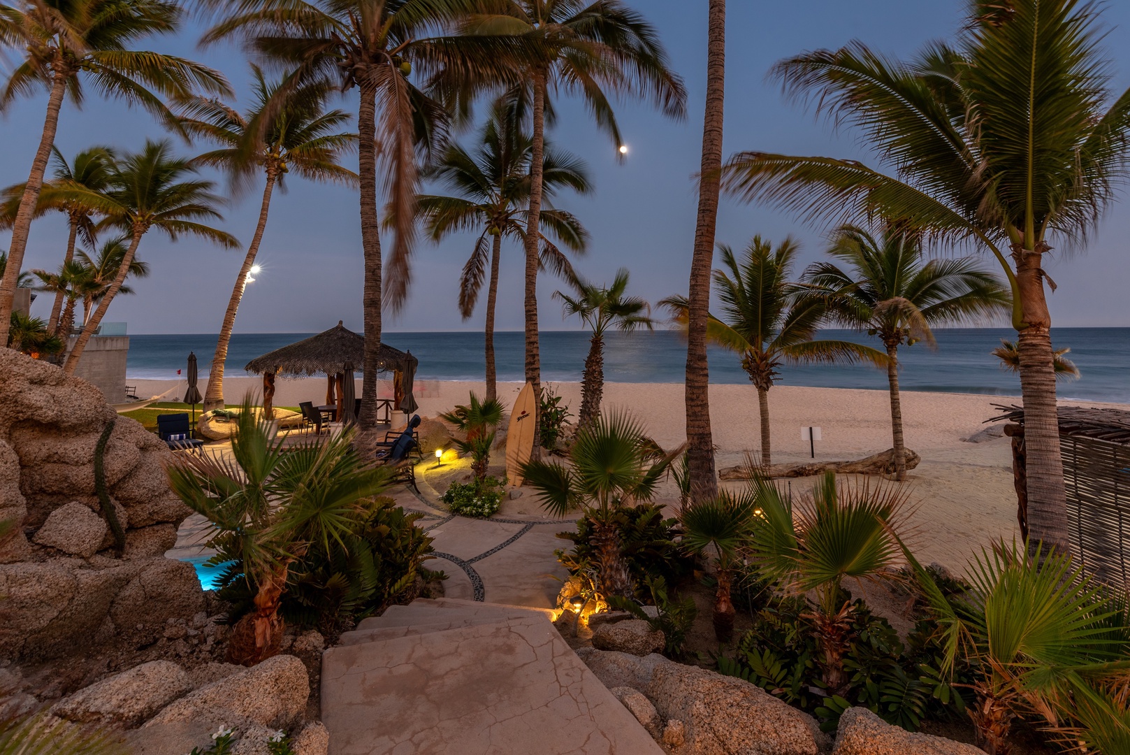 Beachfront property features a thatched palapa, tropical landscaping with palm trees, and ambient lighting along a path leading to sandy shores and calm ocean waters at twilight.